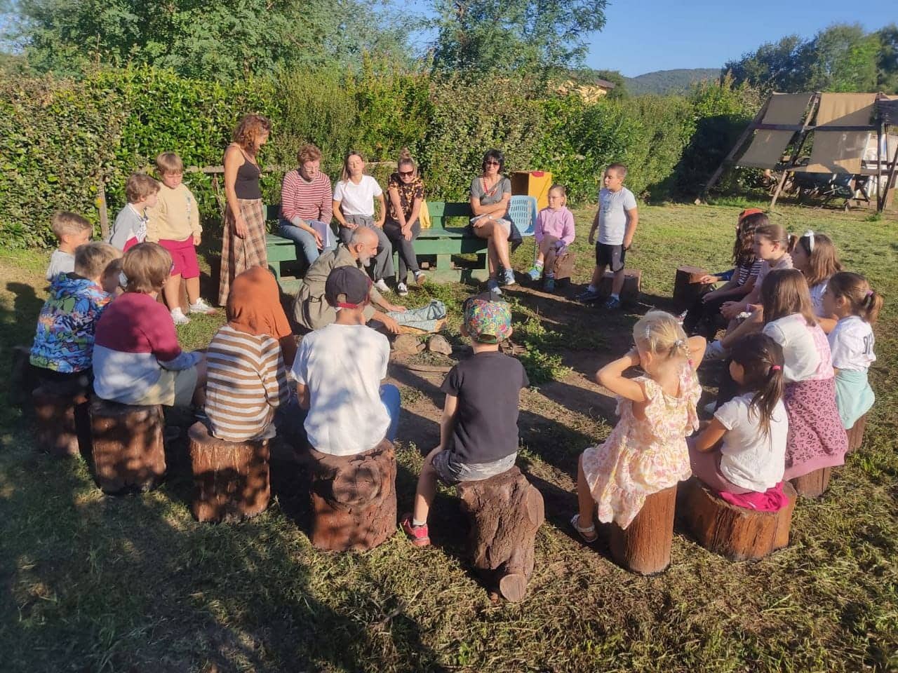 Bambini seduti in cerchio su ceppi di legno in un giardino, con gli educatori